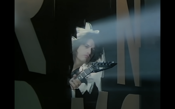 Another still from the video for “Walk This Way”: Steven Tyler of Aerosmith has broken through the wall that separates their rehearsal room from the neighboring studio by Run-DMC. Guitarist Joe Perry takes a peek through the hole where the U once was.