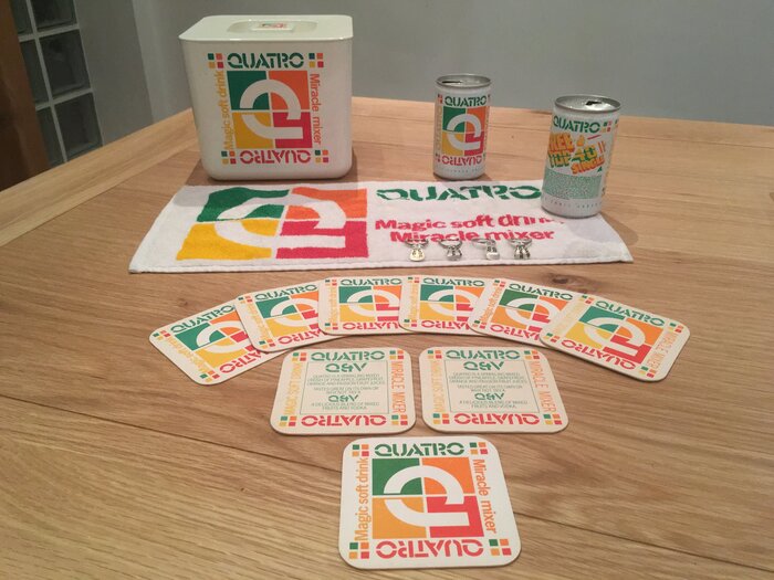 Collection of Quatro paraphernalia incl. coasters, towel, cooling box, from the Quatro Facebook group:
“Quatro is a sparkling mixed crush of pineapple, grapefruit, orange and passion fruit juices. Tastes great on its own or why not try a Q&amp;V, a delicious blend of mixed fruits and vodka / Magic Soft Drink – Miracle Mixer”
The supporting typeface is  or similar.