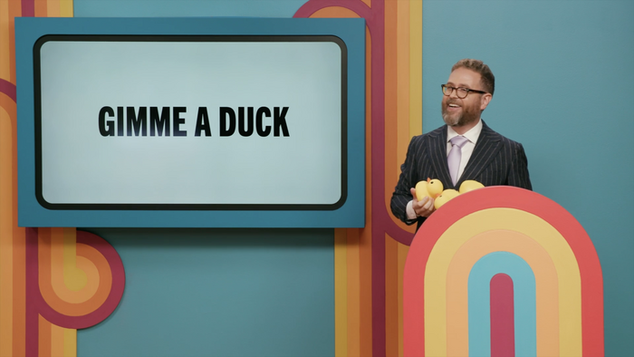 Sam Reich gleefully holding three large rubber ducks behind his podium, with the question prompt displayed on the monitor beside him reading “Gimme a Duck” in Trade Gothic Next
