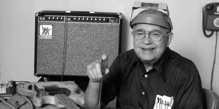 Leo Fender at his CLF Research workshop with a Music Man amp in the background and a Music Man guitar on the table.