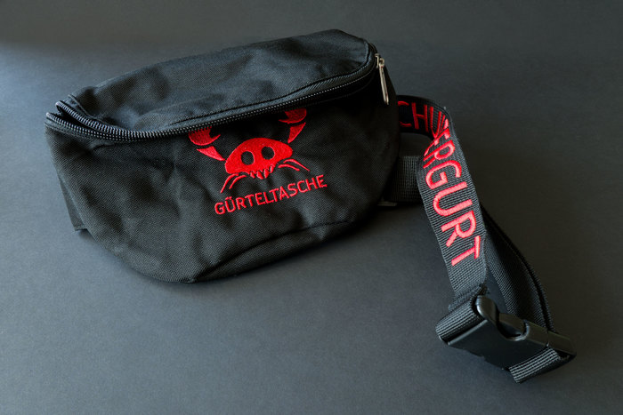 The print on the font of the fanny pack is the German translation of belt pouch – GÜRTELTASCHE. The belt itself reads SCHULTERGURT, German for shoulder-belt.